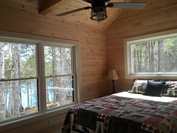 Une chambre de style cabane avec un lit, deux fenêtres, un ventilateur de plafond et une vue sur une forêt et un lac.