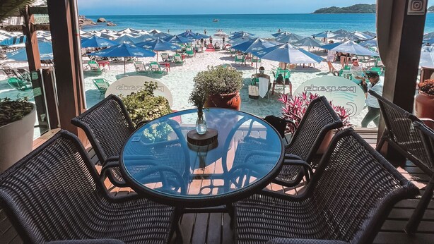 Een overdekt terras met een glazen tafel en rieten stoelen, met uitzicht op een strand met parasols en mensen.