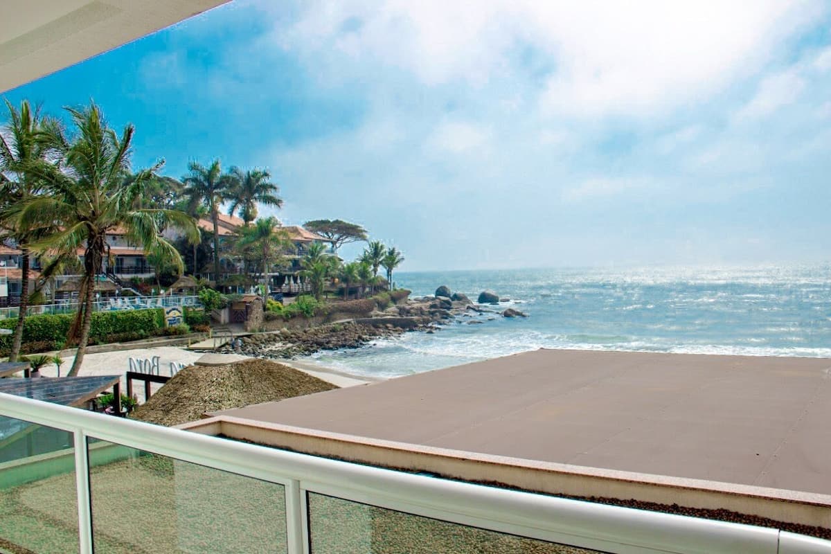 Vista de la playa con palmeras, olas del océano y edificios al fondo.