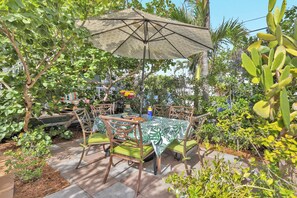 Eine Terrasse im Freien mit Tisch und Stühlen, umgeben von üppiger Vegetation und einem gemusterten Sonnenschirm.