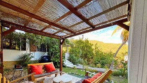 Un'area salotto esterna riparata, con mobili in legno e vista su un giardino e una scalinata che conduce a un edificio.