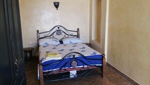 Een bed met een metalen frame en een bedsprei met een patroon, een klein nachtkastje en een wandlamp.