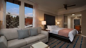 Ein Hotelzimmer mit Bett, Sofa, einem kleinen Tisch und Blick auf Bäume durch große Fenster.