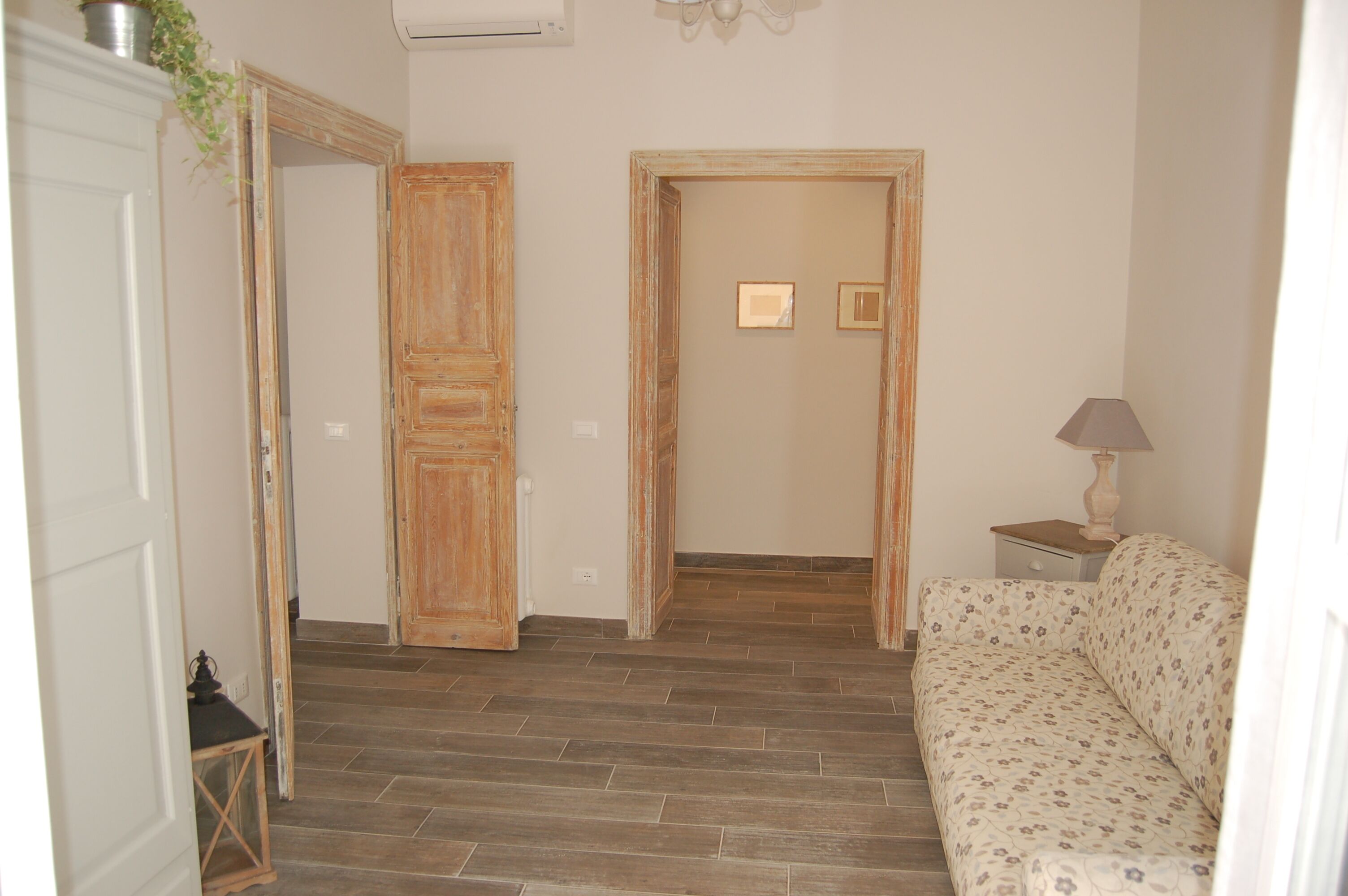 A room with a wooden floor, a sofa, a lamp, and a door leading to another room.