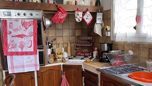 A kitchen with wooden cabinets, a stove, and various kitchen utensils. There is a window with curtains and a shelf with bottles and jars. - for rent Aignan Gers (French Tuscan) (Aignan)