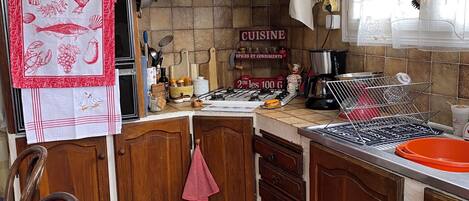 A kitchen with wooden cabinets, a stove, and various kitchen utensils. There is a window with curtains and a shelf with bottles and jars.