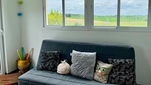 A room with a dark blue sofa, a large window with a view of a green field, and a string of colorful paper decorations. - for rent Aignan Gers (French Tuscan) (Aignan)