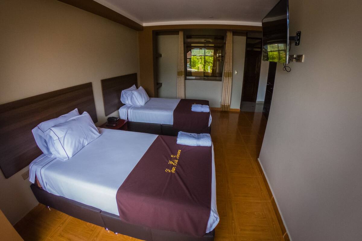 A hotel room with two beds, wooden flooring, a large window with curtains, and a flat-screen TV mounted on the wall.