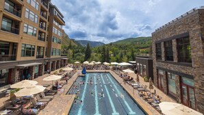 Ein Schwimmbeckenbereich mit mehreren Bahnen, Liegestühlen und Sonnenschirmen, umgeben von Gebäuden mit Balkonen und großen Fenstern.