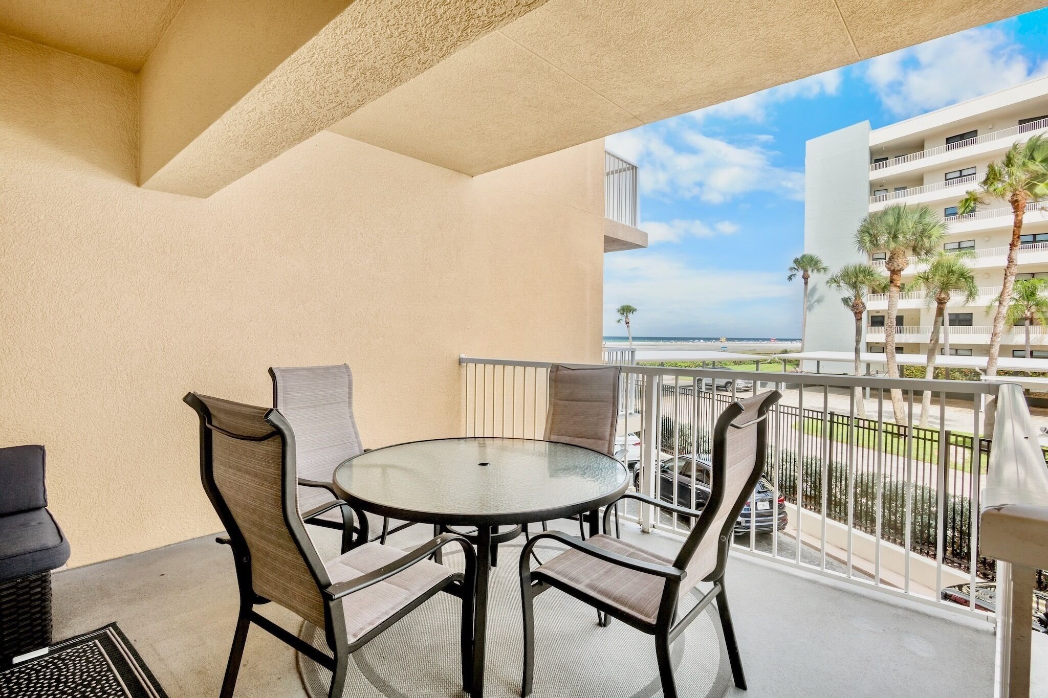 A balcony with a round table and four chairs, overlooking a beach and a hotel building.