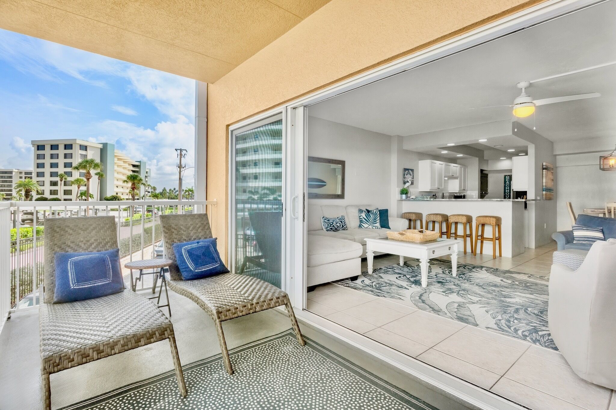 A balcony with wicker furniture, a sliding glass door leading to a living area with a sofa, a coffee table, and a kitchen area with bar stools.