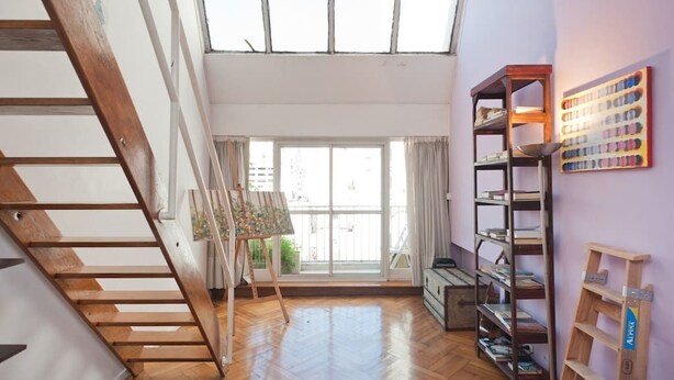 A spacious room with a wooden staircase, a large window with a view, and a bookshelf. - Historico Duplex en el Centro (Buenos Aires)