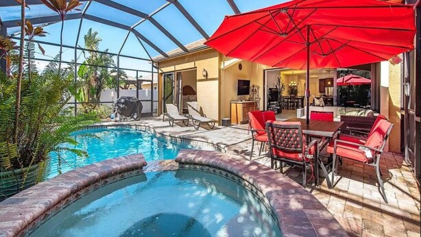 Una piscina interior con techo de vidrio, rodeada de sillones de descanso y un área de comedor con sombrillas rojas.