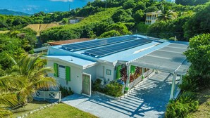 A modern building with solar panels on the roof, surrounded by lush greenery and a clear view of the ocean.