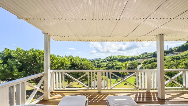 A covered outdoor seating area with two white cushioned chairs and a wooden deck, overlooking a lush green landscape with trees and hills.