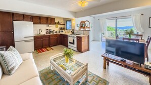 A compact living space with a kitchenette, a sofa, a coffee table with a plant, and a TV on a wooden stand.
