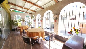 An indoor dining area with round tables and chairs, large arched windows, and potted plants. - Hotel San Felipe Suites (Arequipa)