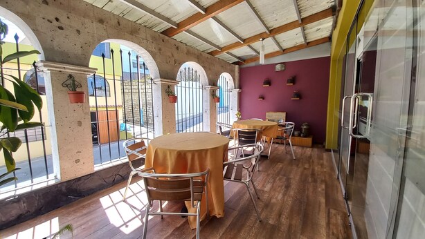 A covered patio with wooden flooring, round tables, and chairs. There are arched windows with metal bars and potted plants. - Hotel San Felipe Suites (Arequipa)
