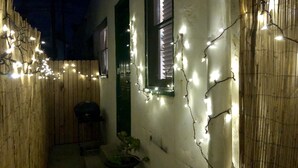 A narrow outdoor corridor with a wooden fence, a potted plant, and string lights. - Bungulow on the beach (Long beacj)