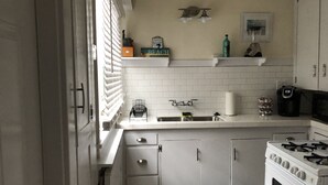 A kitchen with white cabinets, a stove, a sink, and a window with blinds. - Bungulow on the beach (Long beacj)