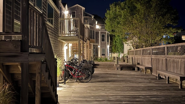 Une promenade en bois mène à une rangée de maisons dotées de balcons. Un vélo est garé sur la promenade, et un arbre se trouve à droite.