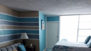 Une chambre avec un canapé gris, un lit recouvert d’une couette à motifs floraux, une petite table avec une lampe, et une vue sur l’extérieur par une fenêtre.