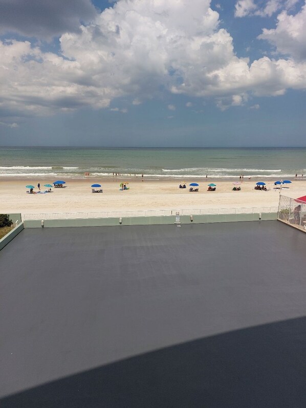 Vista a la playa desde un balcón, con cielo despejado, arena y varias sombrillas.