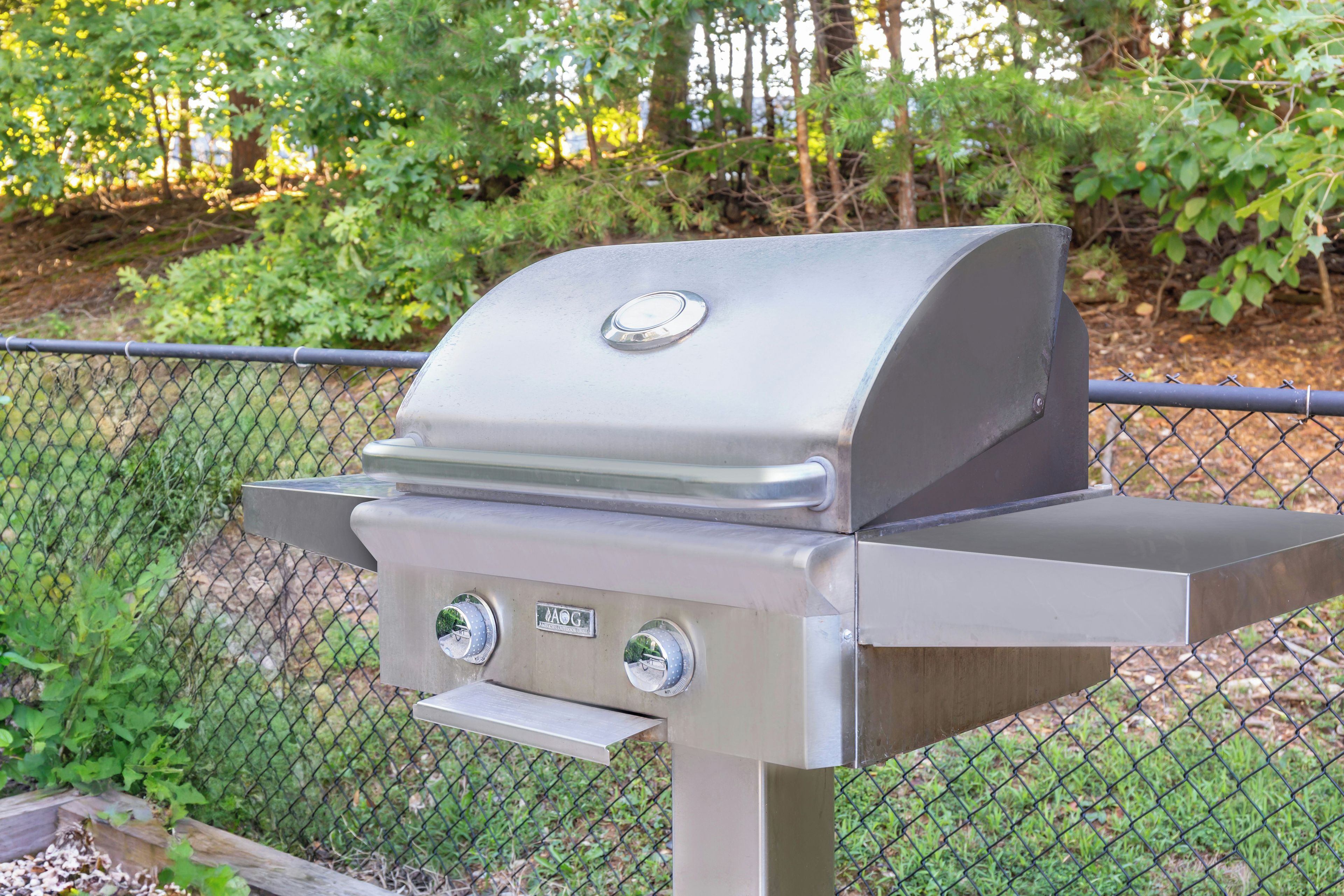 A stainless steel barbecue grill with a digital thermometer and control knobs, mounted on a metal stand with a chain-link fence in the background.