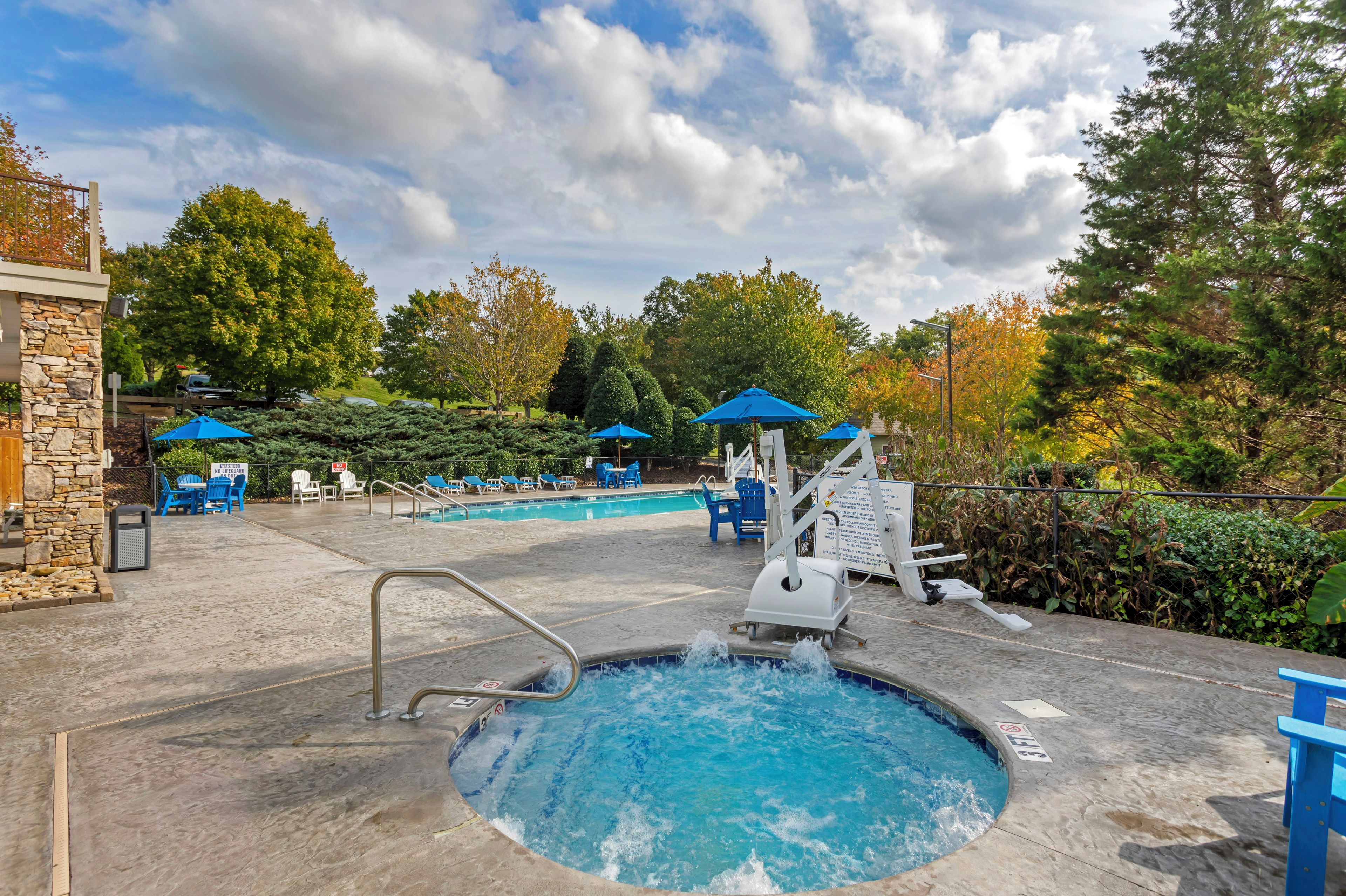 A swimming pool with a hot tub, surrounded by lounge chairs and umbrellas, with trees and a stone wall in the background.