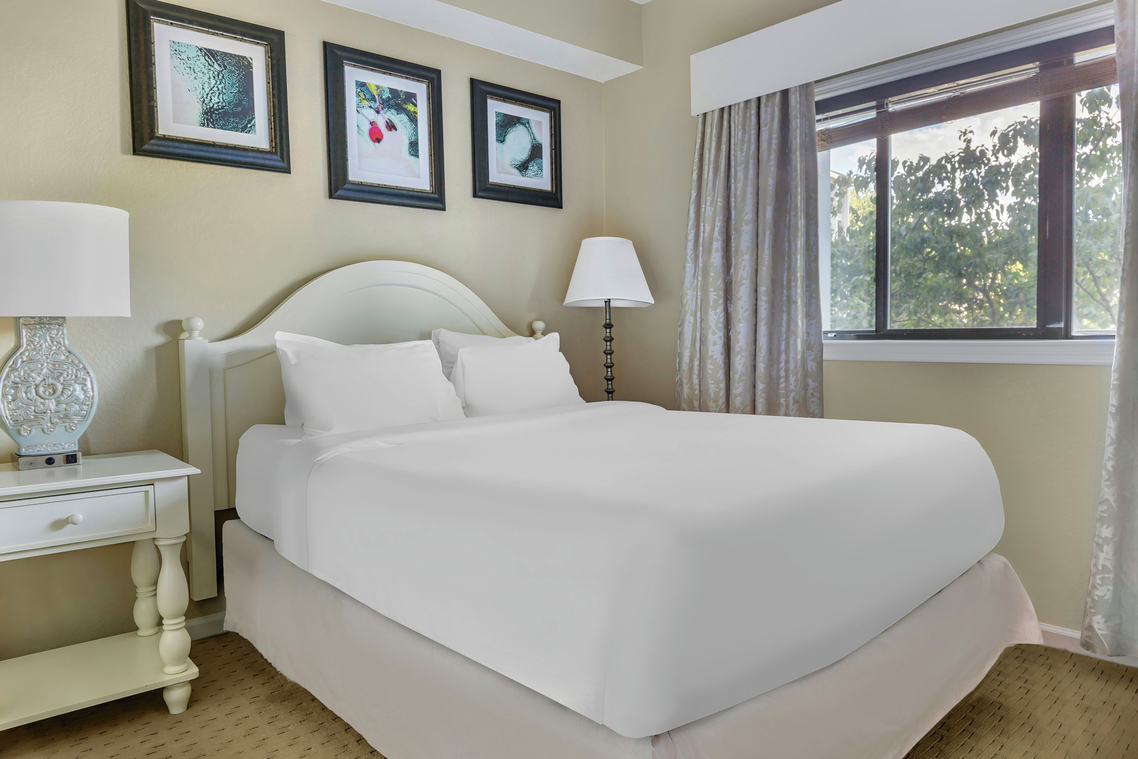 Habitación de hotel con una cama grande, dos mesitas de noche, un ventanal con cortinas y tres cuadros enmarcados en la pared.