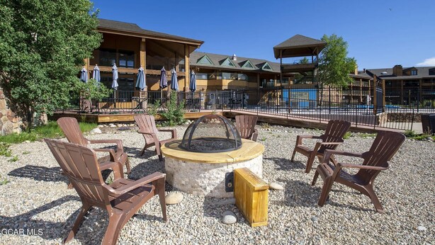 A fire pit with chairs and a wooden box, surrounded by gravel, with a building in the background.