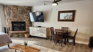 A cozy living room with a stone fireplace, a flat-screen TV mounted on the wall, a wooden dining table with chairs, and a large window allowing natural light.