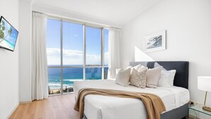 Een slaapkamer met een groot bed, uitzicht op de oceaan, een ingelijst schilderij aan de muur en een bedlampje.