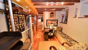 Un salón acogedor con un sofá, un escritorio y una silla. Tiene techo de vigas de madera y una estantería montada en la pared con objetos decorativos.