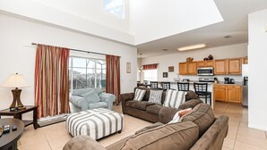 Ein geräumiges Wohnzimmer mit einer Couch, einem Hocker und einem Essbereich, im Hintergrund ist eine Küche erkennbar.