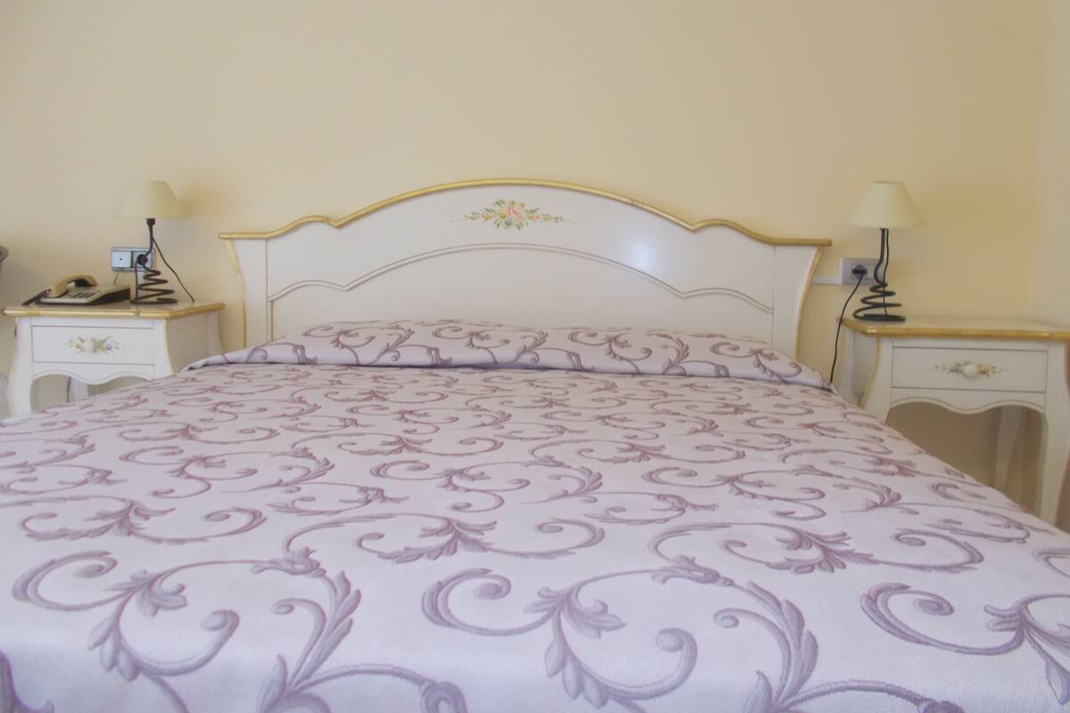 Un lit bien fait, avec une tête de lit à motifs floraux et une couvre-lit à dessins. On y trouve deux tables de chevet, chacune agrémentée d’une lampe, et un téléphone posé sur l’une d’elles.