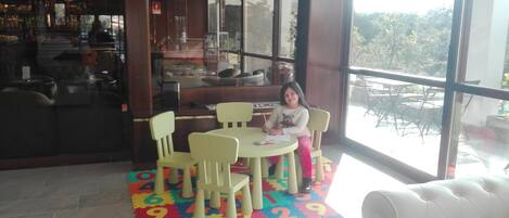 Niño sentado a una mesa con un tapete de juego de colores, una ventana con vistas a muebles de exterior, y una zona de bar al fondo.