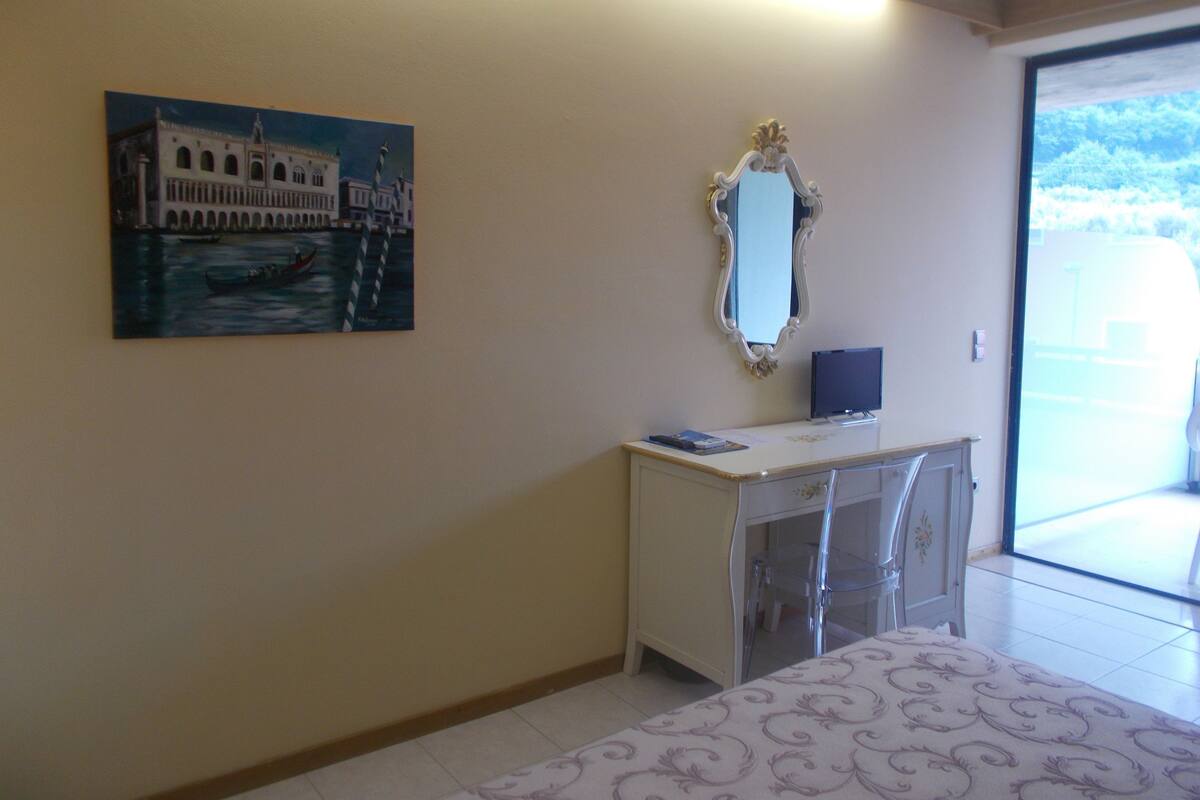 Une chambre d’hôtel avec un bureau, un miroir, un tableau représentant un pont et une vue sur les arbres par la fenêtre.