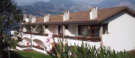 A modern two-story house with a balcony, surrounded by lush greenery and mountains in the background.