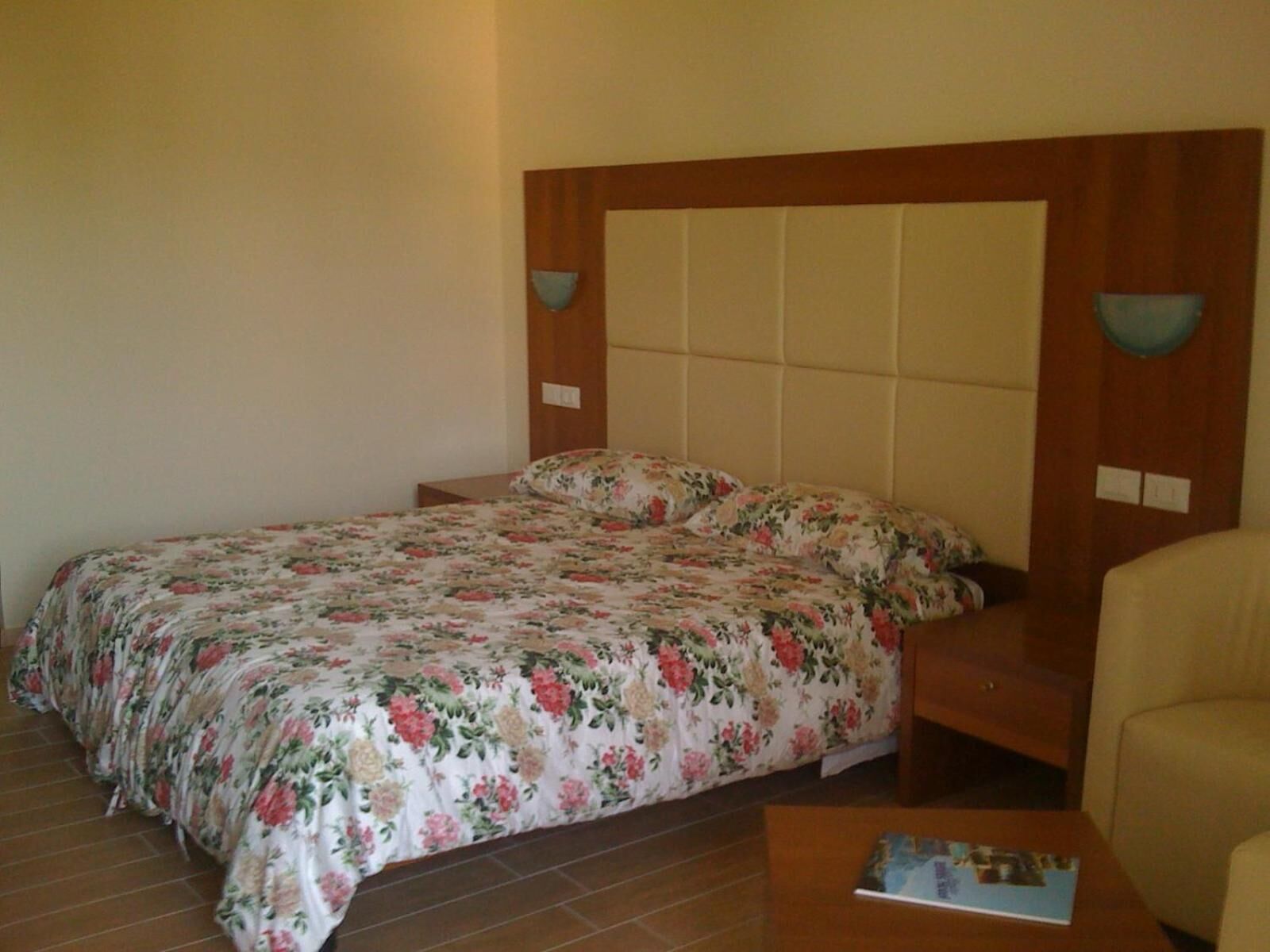 Een slaapkamer met een bed, een houten hoofdbord, een nachtkastje, een stoel en een klein tafeltje met een tijdschrift.