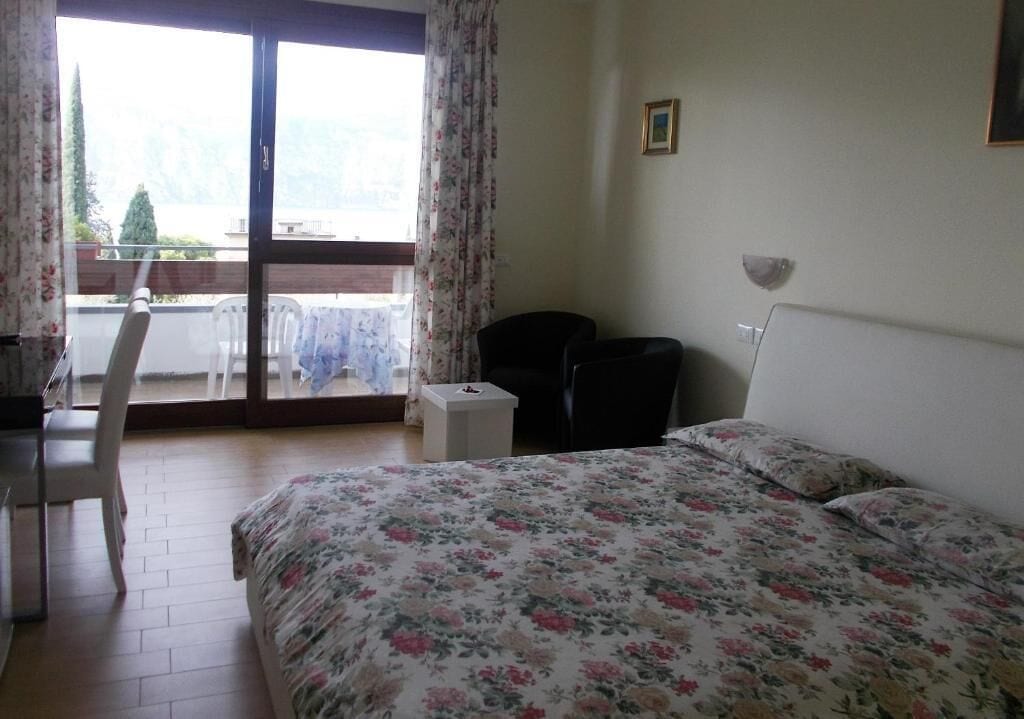 Quarto com cama, escrivaninha e cadeira. Há uma janela com cortinas e é visível uma varanda lá fora.