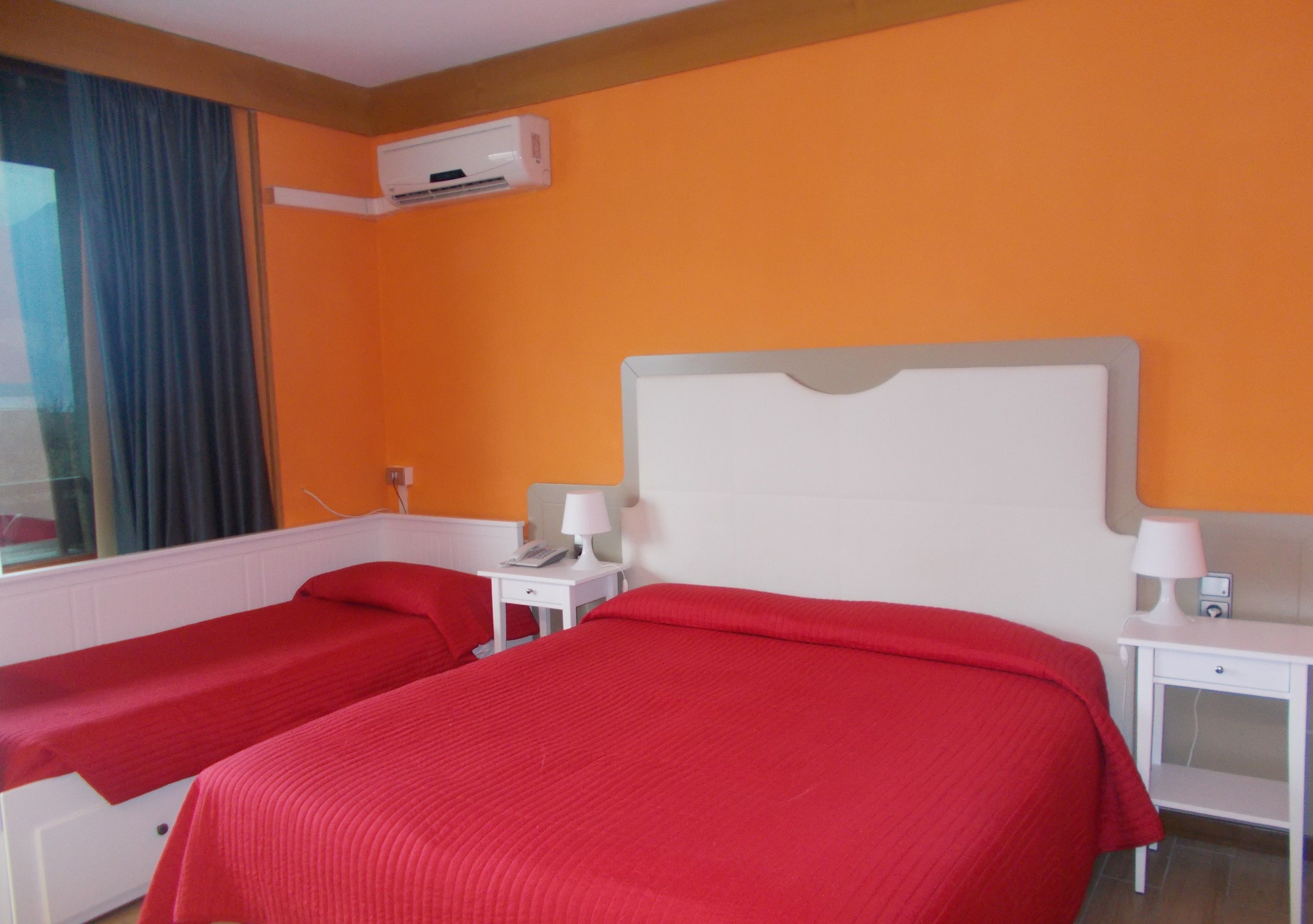 A hotel room with two beds, an air conditioning unit, a window with curtains, and a bedside table with a lamp and a phone.