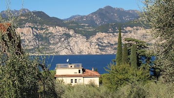 Un bâtiment coiffé d’un toit de tuiles rouges, entouré d’oliviers et de cyprès, qui surplombe une étendue d’eau avec des montagnes en arrière-plan.