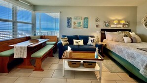 A living room with a sofa, a bench, a table, and a bed. There are windows with blinds, wall art, and a ceiling light. - The Sea Star is an adorable seaside studio with a gorgeous ocean view! (Daytona Beach)