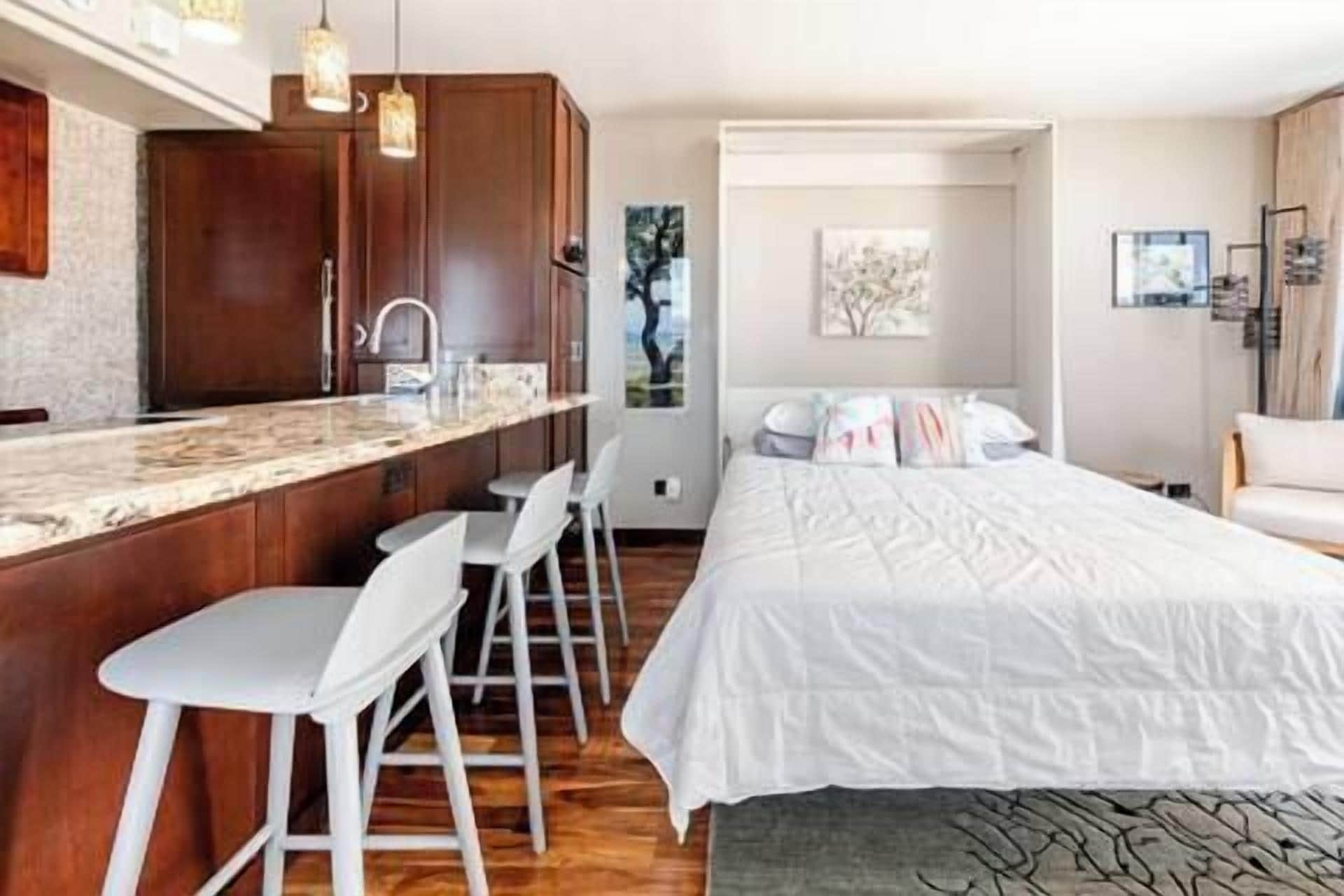 A hotel room with a kitchen area, a bed with white bedding, and a seating area with white chairs.