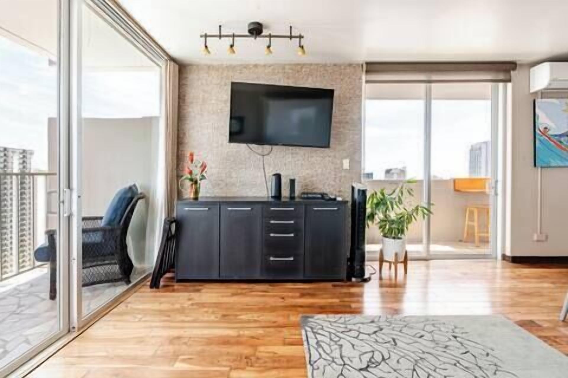 A modern living room with a flat-screen TV, a dark wooden cabinet, and a sliding glass door leading to a balcony.