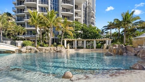 A clear pool with rocks, palm trees, and a building with balconies.