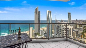 A balcony with a table, a bottle, and glasses, overlooking a beach and cityscape.