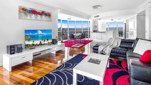 A modern living room with a flat-screen TV, a white entertainment unit, a black leather sofa, a red rug, and a city view through large windows.