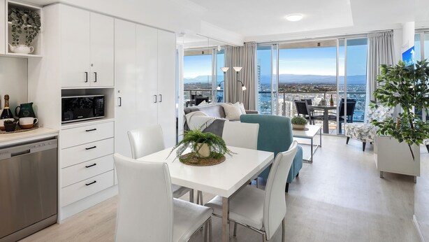 A modern kitchen with white cabinets, a dishwasher, and a microwave. There is a dining area with a white table and chairs, and a balcony with a view of the city. - Chevron Renaissance River View (Surfers Paradise)